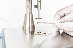Close-up of a hand holding a pen dispensing thick liquid beneath a piece of laboratory equipment.